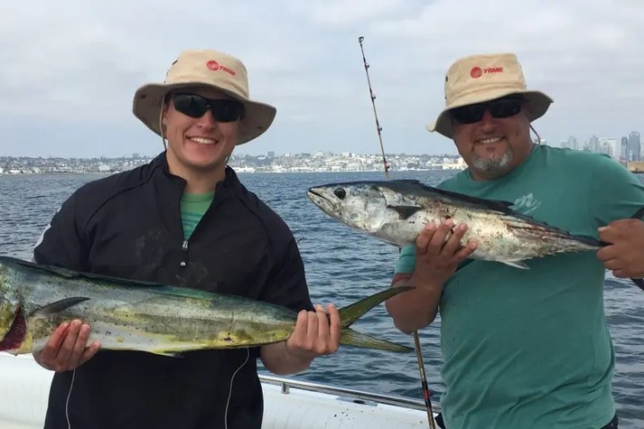 Two men on a boat holding large fish, wearing hats and sunglasses, with a cityscape in the background.