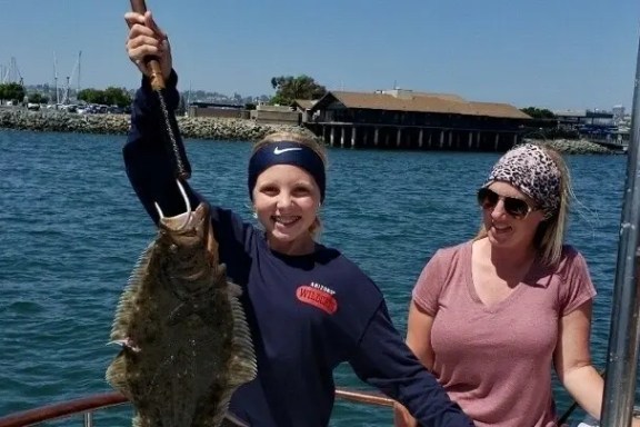 Two people on a boat, one holding a large fish against a waterfront backdrop.
