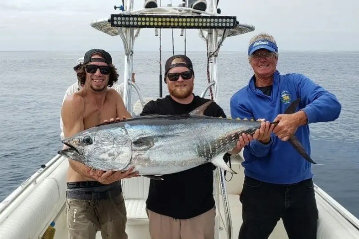Three people on a boat holding a large tuna fish against an ocean backdrop.