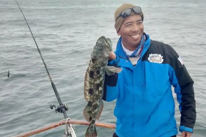 Smiling person in a blue jacket holds a fish on a boat in open water.
