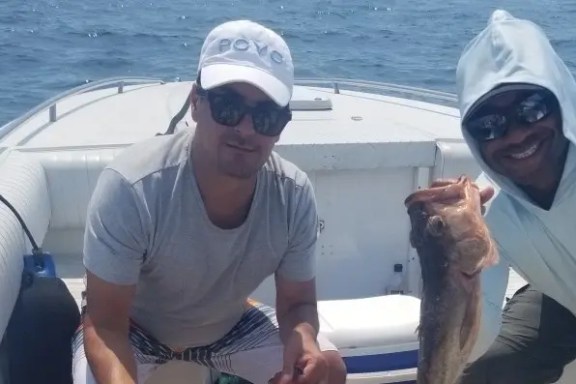 Two men on a boat holding a fish, smiling toward the camera.
