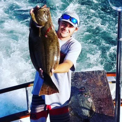 Person in cap and sunglasses holds a large fish on a boat with water in the background.
