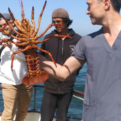 Man holding large lobster on a boat with two others nearby, ocean in the background.