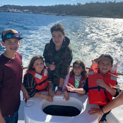 Five kids on a boat wearing life jackets, smiling with a lake and hills in the background.