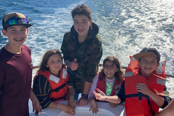 Five kids on a boat wearing life jackets, smiling with a lake and hills in the background.