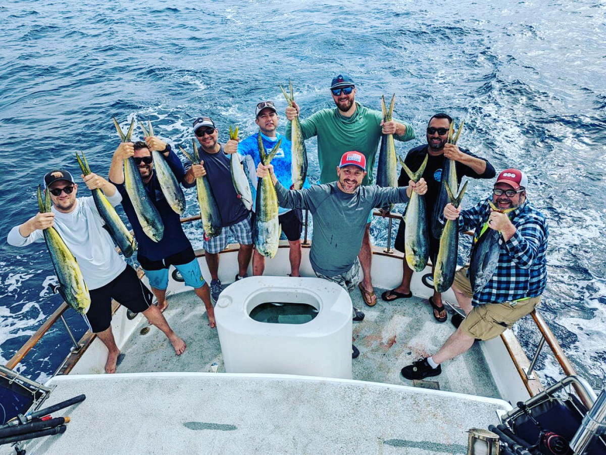 Eight men on a boat holding freshly caught fish over the ocean.